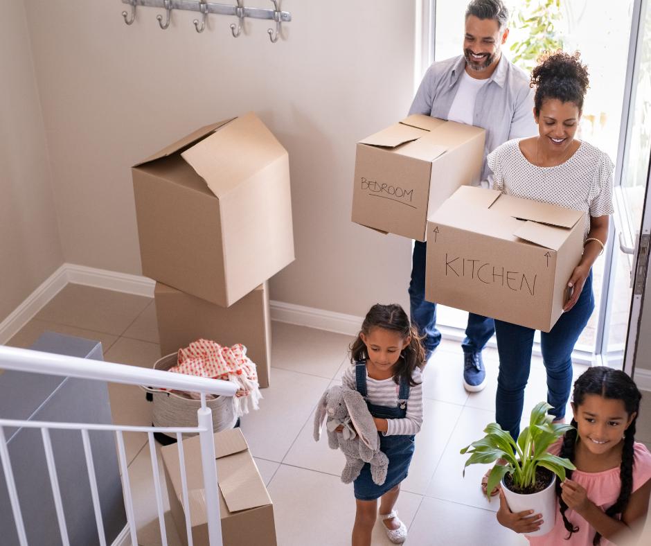 A family moving into a new home carrying cardboard boxes. There are two children and a woman and a man. the man is carrying a box which says bedroom and the woman is carrying a box which says kitchen.