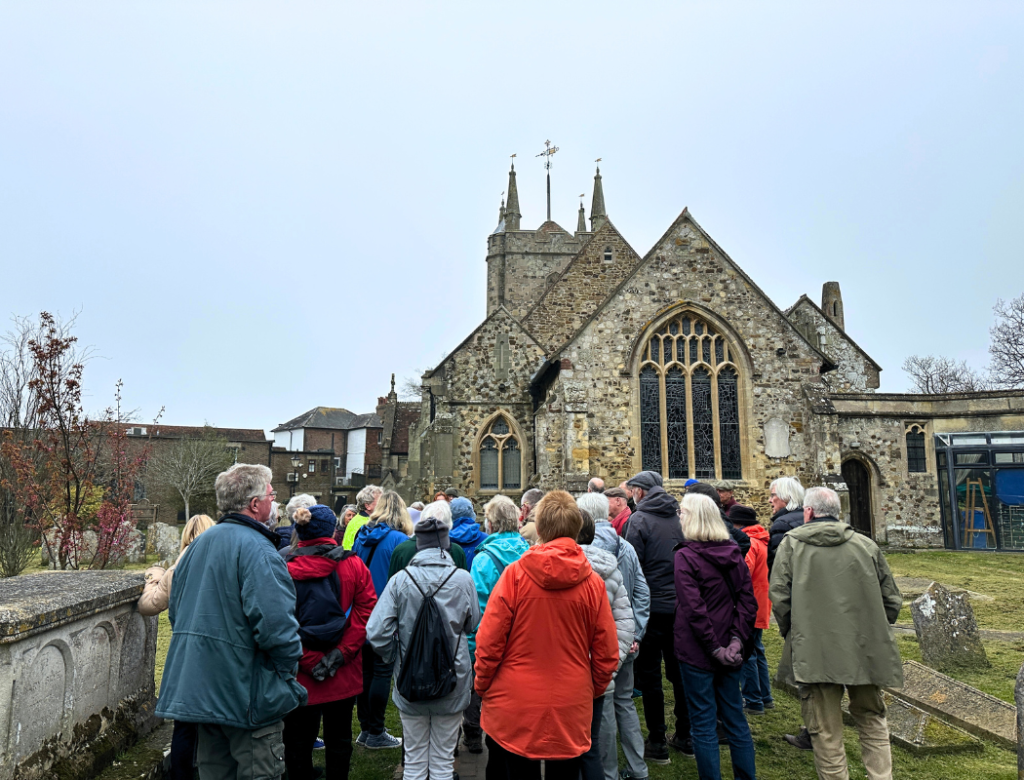 Large group of walkers stood listening to Graham talk in Hailsham graveyard, with church behind them.