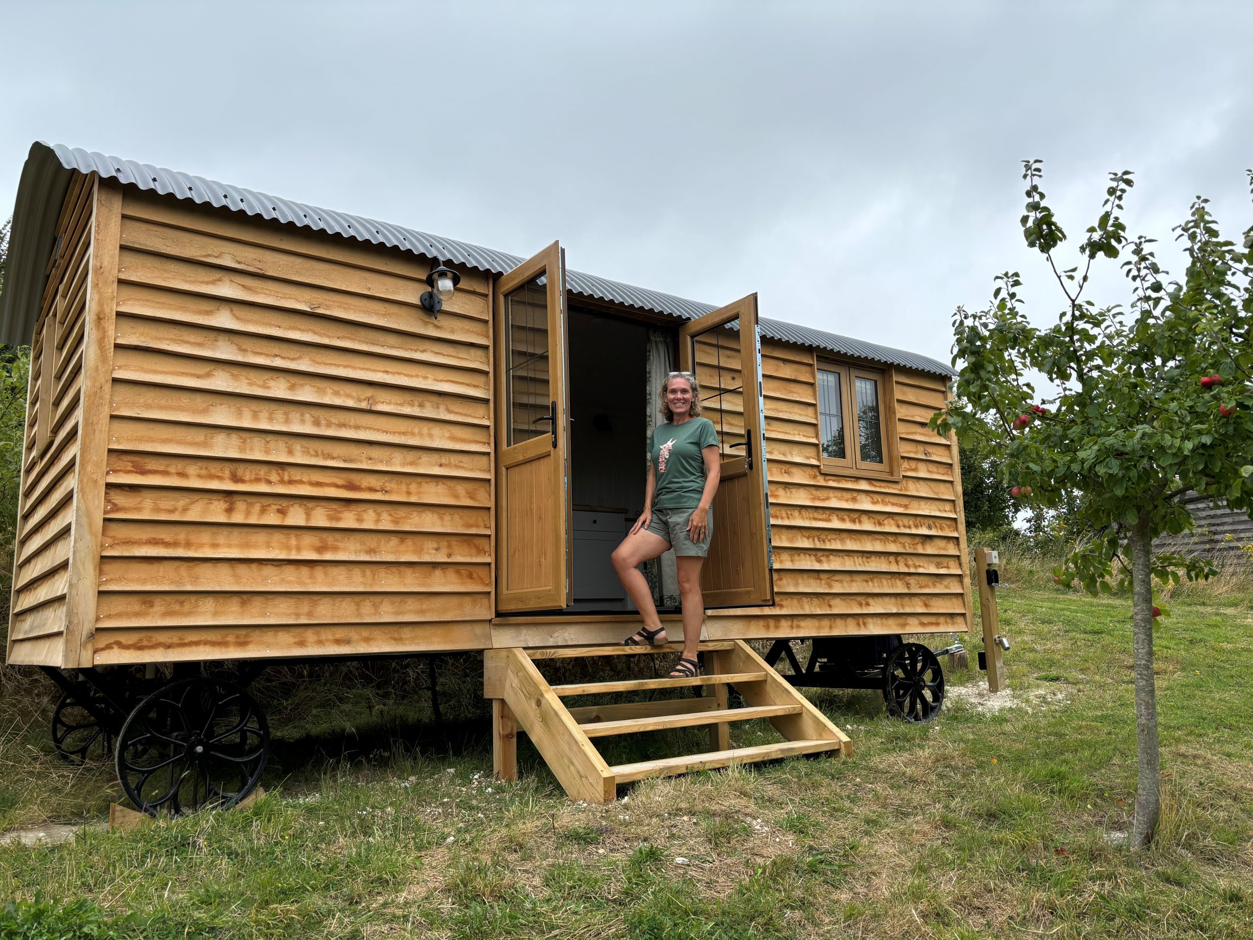 Wooden cabin on grass with wooden steps leading up to it. There is a lady wearing a pair of shorts and green t.shirt standing on the steps looking towards the camera